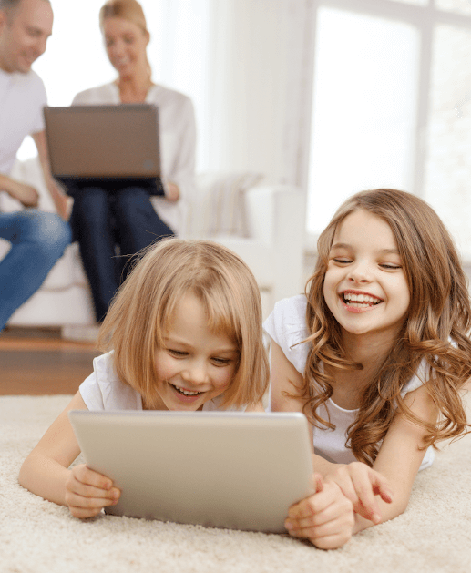 Zdjęcie przedstawia dwie śmiejące się dziewczynki trzymające i spoglądające w tablet. Za dziećmi widać uśmiechniętych rodziców korzystających z laptopa.